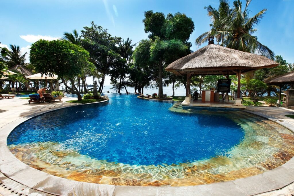 Outdoor infinity swimming pool surrounded by trees and a thatched gazebo, overlooking the sea